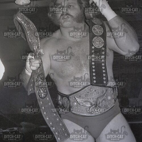 Professional wrestler posing shirtless with two championship belts, one draped over shoulder and the other around the waist.