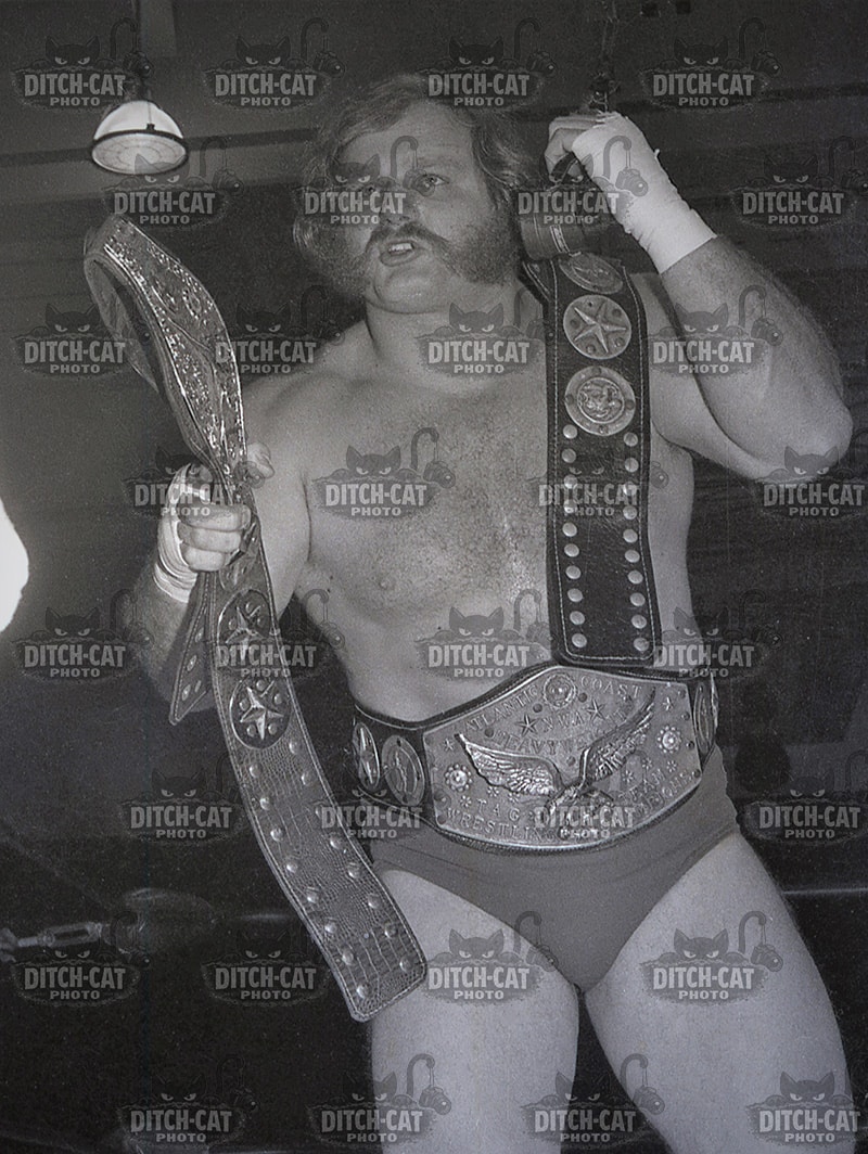 Professional wrestler posing shirtless with two championship belts, one draped over shoulder and the other around the waist.