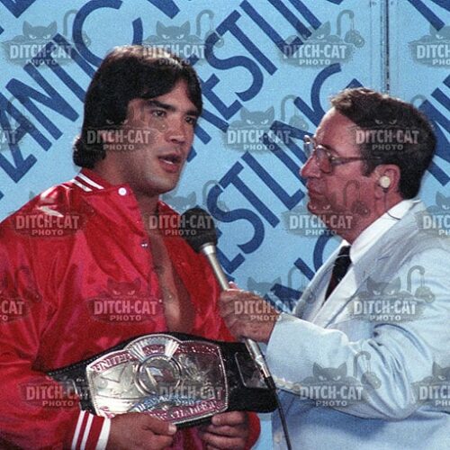 Wrestler in a red jacket holding a championship belt is interviewed by a reporter with a microphone, blue backdrop says champion.