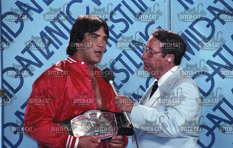 Wrestler in a red jacket holding a championship belt is interviewed by a reporter with a microphone, blue backdrop says champion.