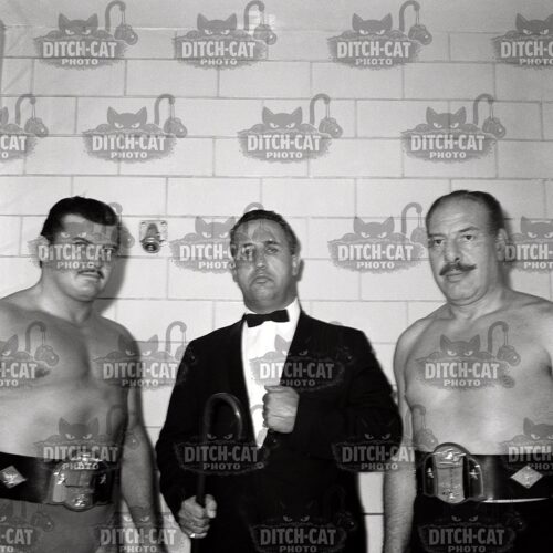 Three men pose for a black-and-white photo: two muscular wrestlers with championship belts stand on either side of a suited promoter, who holds a microphone in front of a tiled wall.