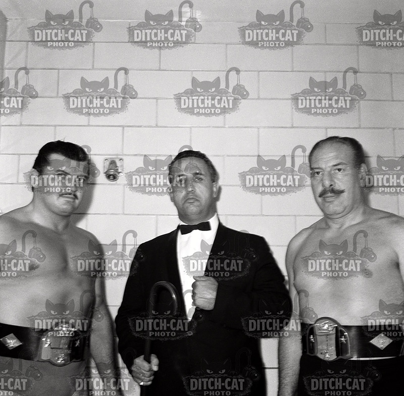 Three men pose for a black-and-white photo: two muscular wrestlers with championship belts stand on either side of a suited promoter, who holds a microphone in front of a tiled wall.