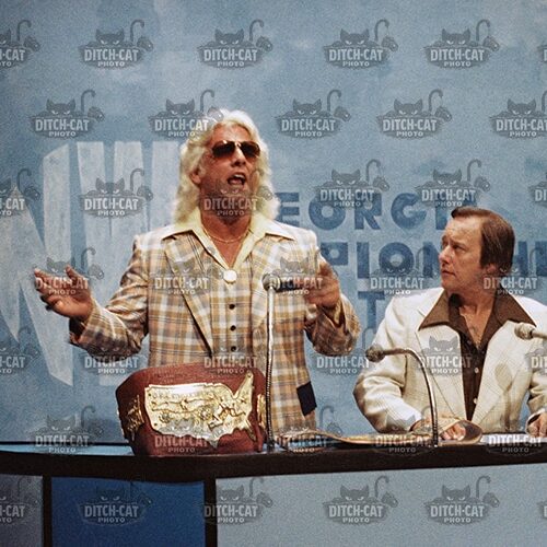 Two men sit behind a TV news desk with microphones; left man with long blond hair and plaid jacket speaks, right man in white suit listens; a decorative bag rests on the desk.