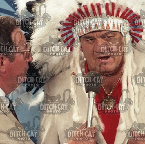 Man wearing a Native American feathered headdress is interviewed on stage, speaking into a microphone beside a suited interviewer.