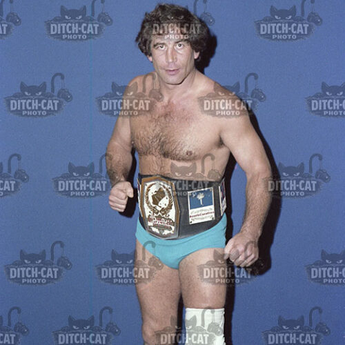 Male professional wrestler posing with a championship belt, wearing blue trunks, boots, and a knee wrap against a blue backdrop.