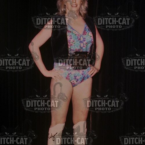 Female performer on stage posing with hands on hips, wearing a colorful leotard, black vest, and white knee-high boots.