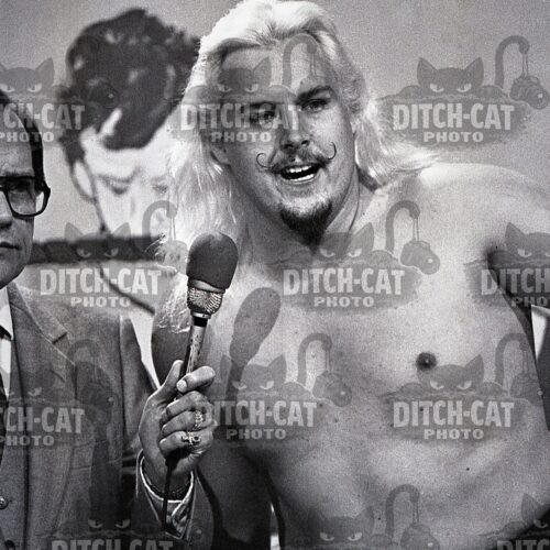 Blond, mustached man shirtless being interviewed at a press event, holding a round Georgia sign; interviewer in a suit beside him.