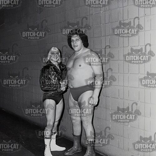 Two men pose together in a backstage hallway; one wears a leather jacket with shorts and white socks, the other is shirtless in briefs with wrestling boots.
