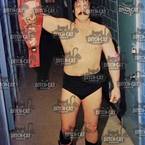 Male wrestler shirtless in a locker room raising a red championship belt overhead in triumph.