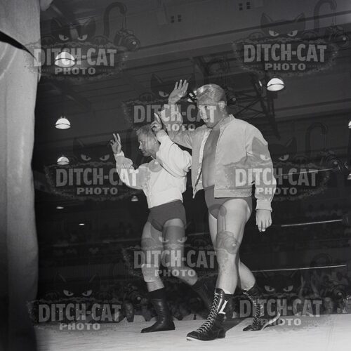 Two male wrestlers in a ring raise their hands in victory, wearing trunks and boots, with a cheering audience in the background.