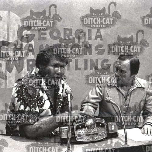 Two men sit at a table with microphones during a press conference; a championship belt rests in front of them, with a branded backdrop in the background.