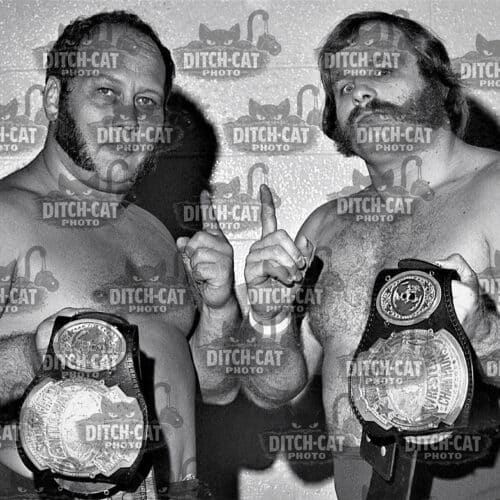 Two muscular wrestlers pose shirtless, each wearing a championship belt over their waists, showing thumbs up.