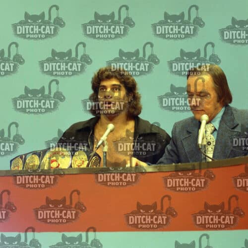 Two people at a press conference speaking into microphones, with a teal backdrop covered in Ditch-Cat Photo logos.